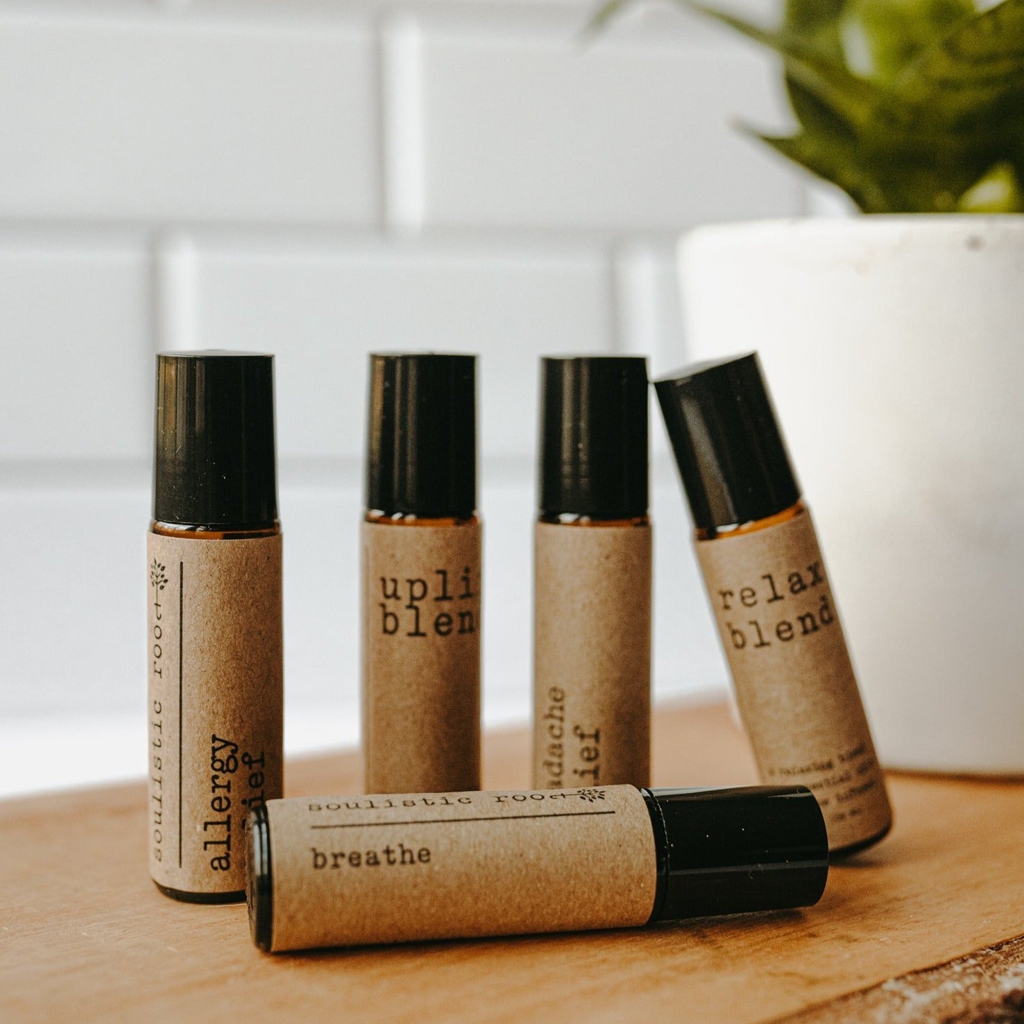 Essential oil rollers. There are five different blends -uplift which has rose quartz gemstones, headache relief, relax blend which has amethyst gemstones, allergy relief and breathe. They are in amber roller bottles on a wood block in front of a plant and subway tiles.