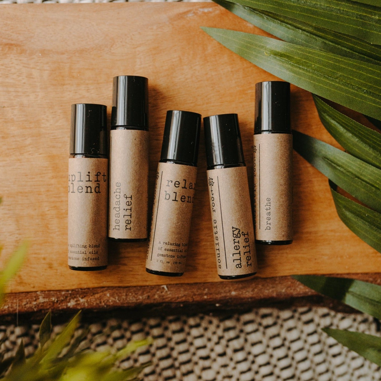 Essential oil rollers. There are five different blends -uplift which has rose quartz gemstones, headache relief, relax blend which has amethyst gemstones, allergy relief and breathe. They are in amber roller bottles on a wood block in next to a palm leaf.